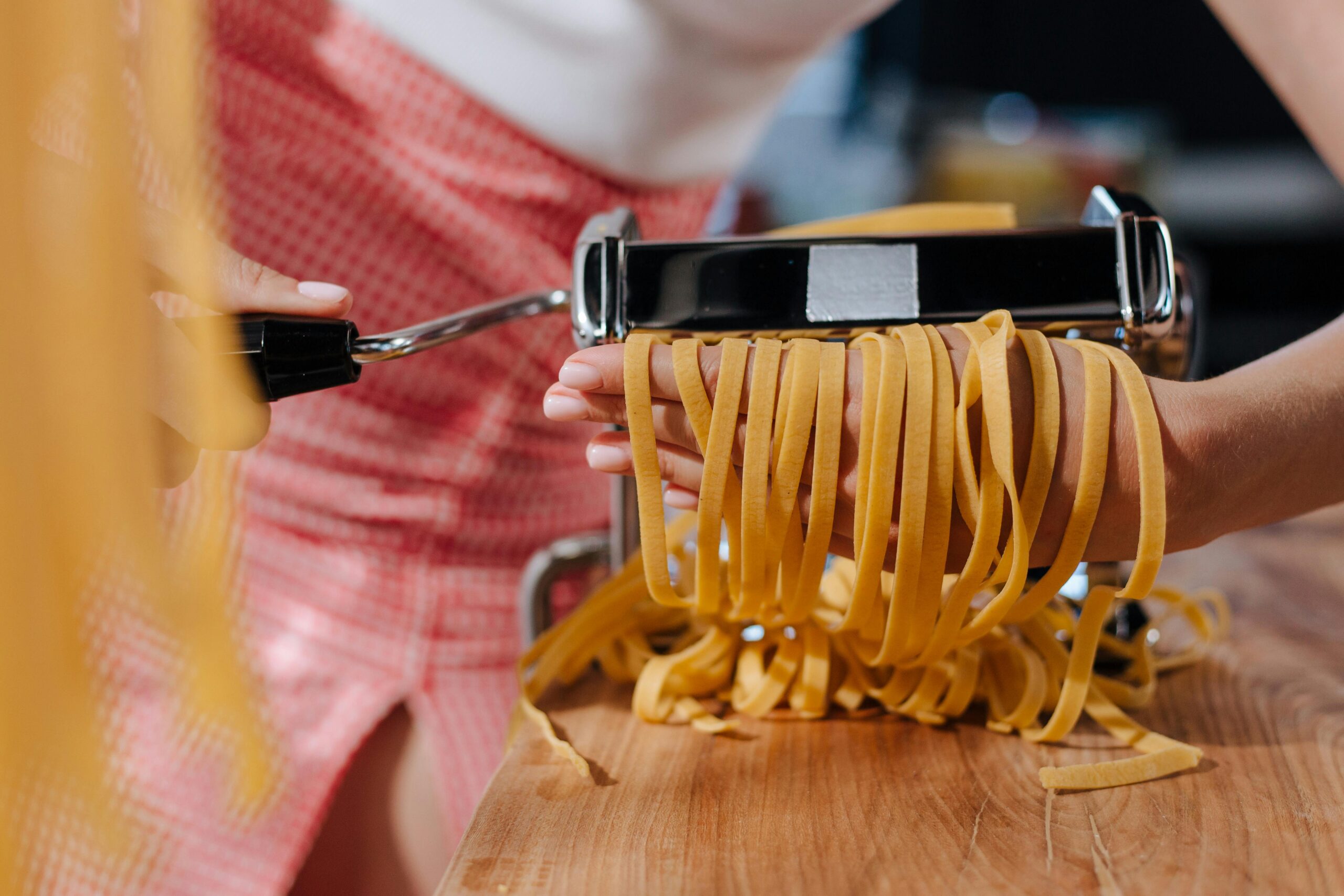 A close-up of hands using a pasta maker, with fresh, yellow fettuccine strands cascading from the machine onto a wooden surface.