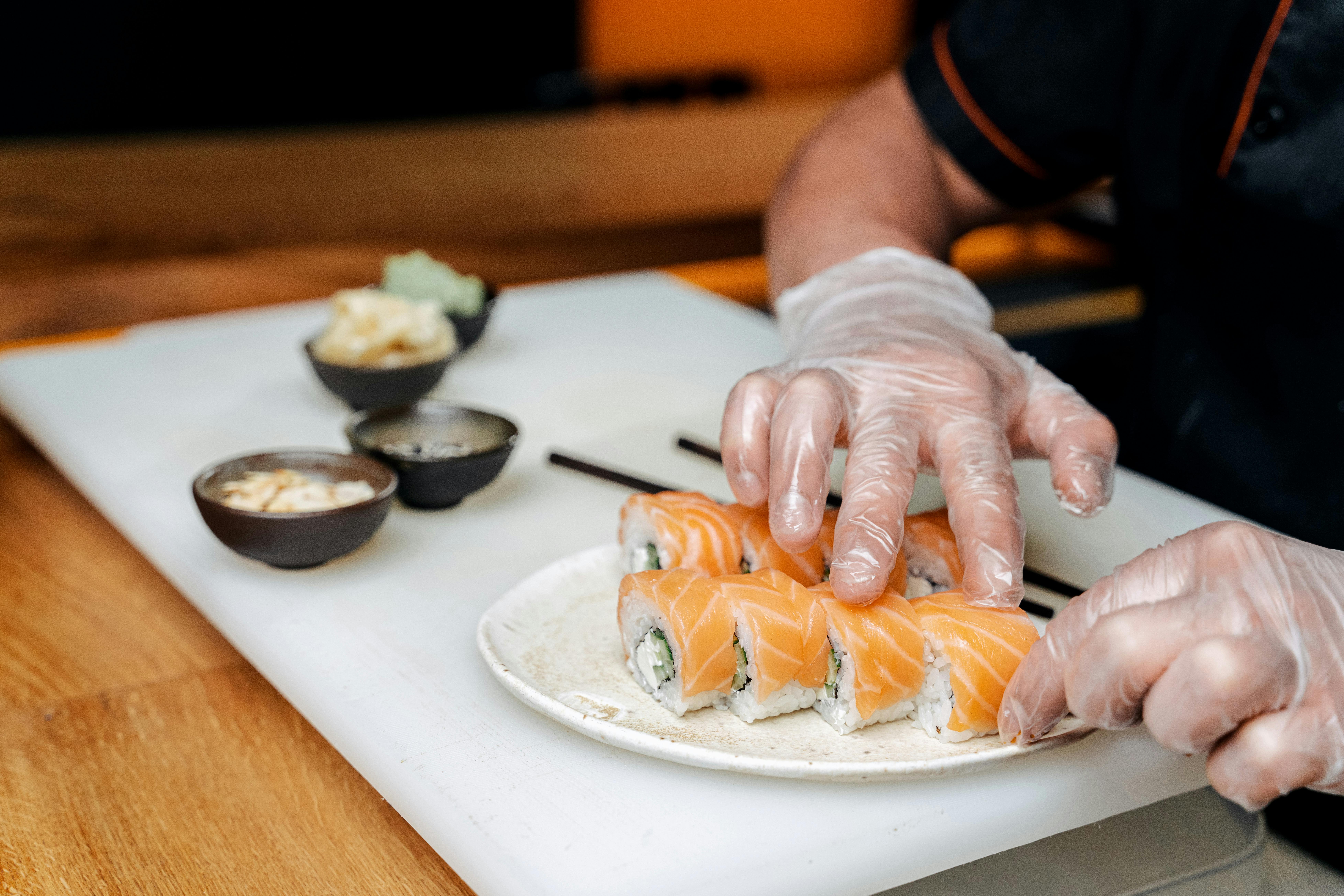 A chef in gloves carefully arranges salmon sushi rolls on a white plate, with small bowls of wasabi and ginger nearby.