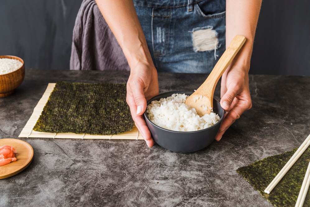 A person holds a bowl of cooked rice, with nori sheets, chopsticks, and pickled ginger on a textured countertop, preparing sushi.