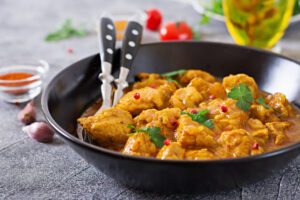 A bowl of flavorful chicken in curry sauce, garnished with cilantro and pink peppercorns, with serving utensils on the side.