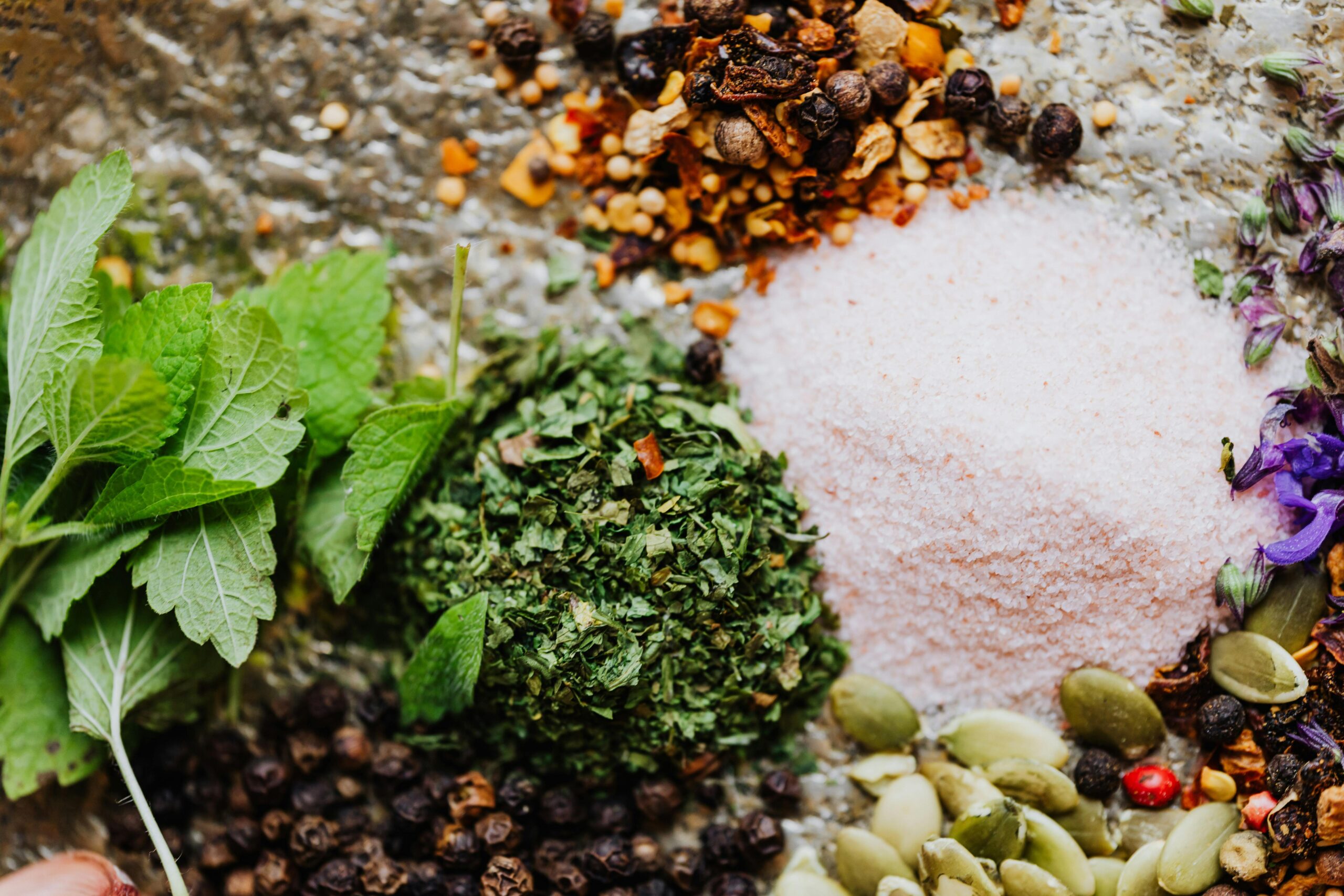 A colorful arrangement of fresh herbs, spices, and pink salt on a textured surface, showcasing vibrant greens, browns, and purples.
