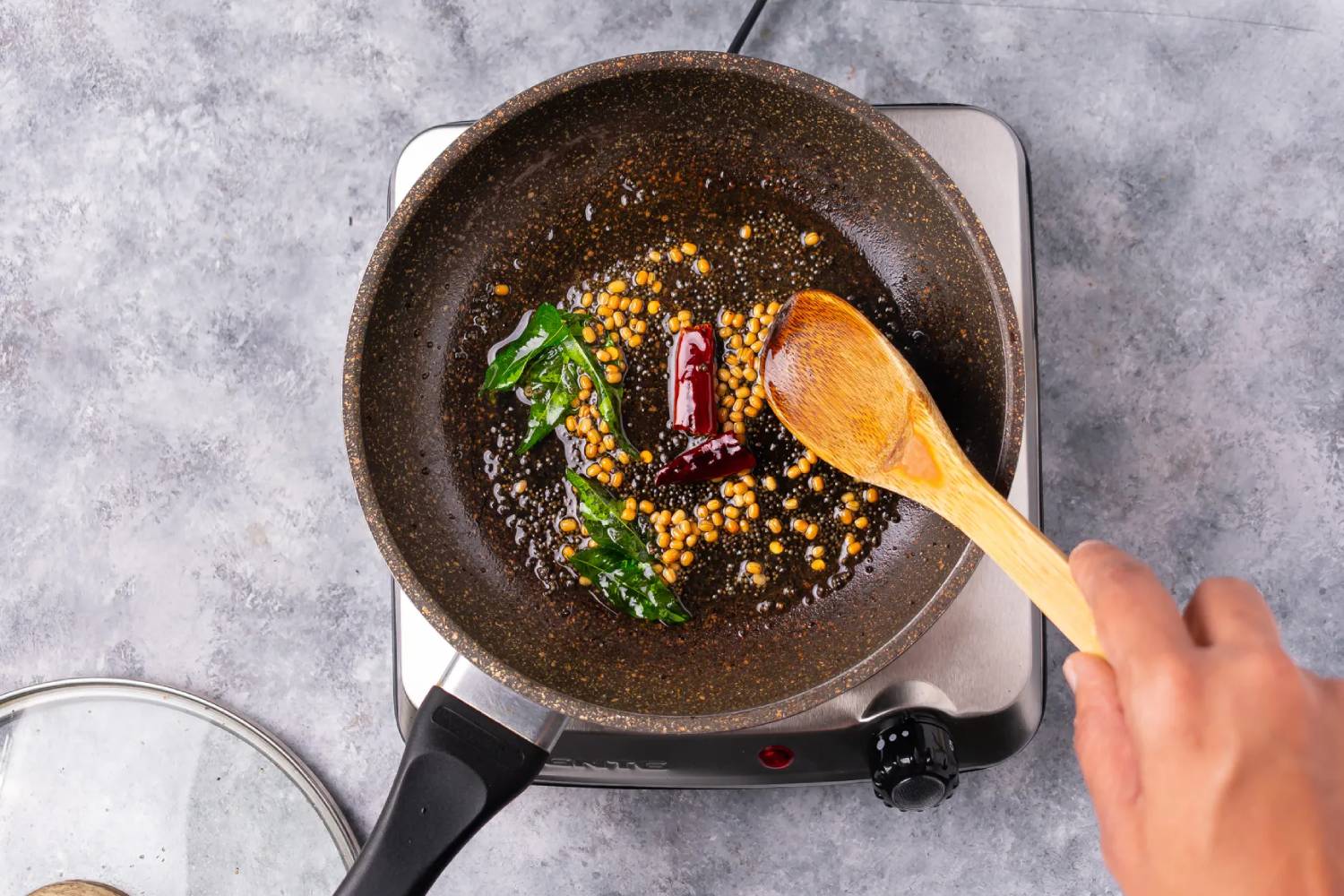 A hand stirs mustard seeds, curry leaves, and dried red chilies in a frying pan on a stovetop, with a wooden spoon.