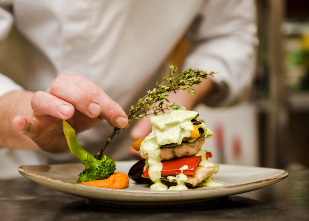 A chef carefully garnishes a layered dish of chicken, vegetables, and sauce with a sprig of thyme, enhancing its presentation.