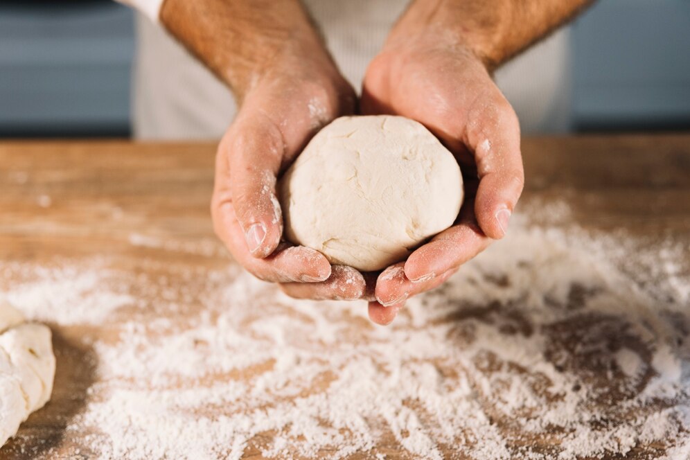 A pair of hands hold a round ball of dough above a wooden surface dusted with flour.