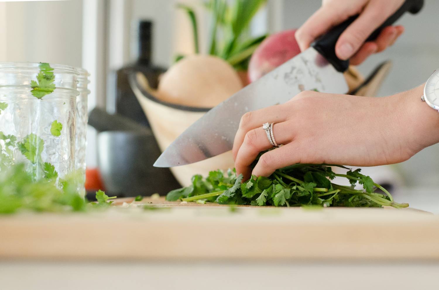 A close-up of a hand slicing fresh cilantro on a wooden cutting board, with a knife and kitchen items in the background.