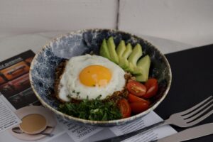 Bowl of buckwheat topped with a fried egg, fresh tomatoes, avocado slices, and sprinkled with herbs, resting on a magazine backdrop.