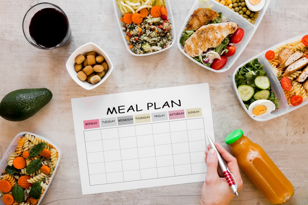 A meal plan sheet surrounded by healthy meals, snacks, and beverages, with a hand holding a pen, ready to fill in the plan.