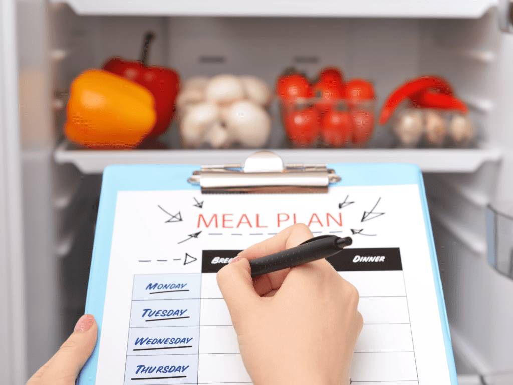 A person writing a meal plan on a clipboard in front of a refrigerator filled with colorful vegetables and eggs.