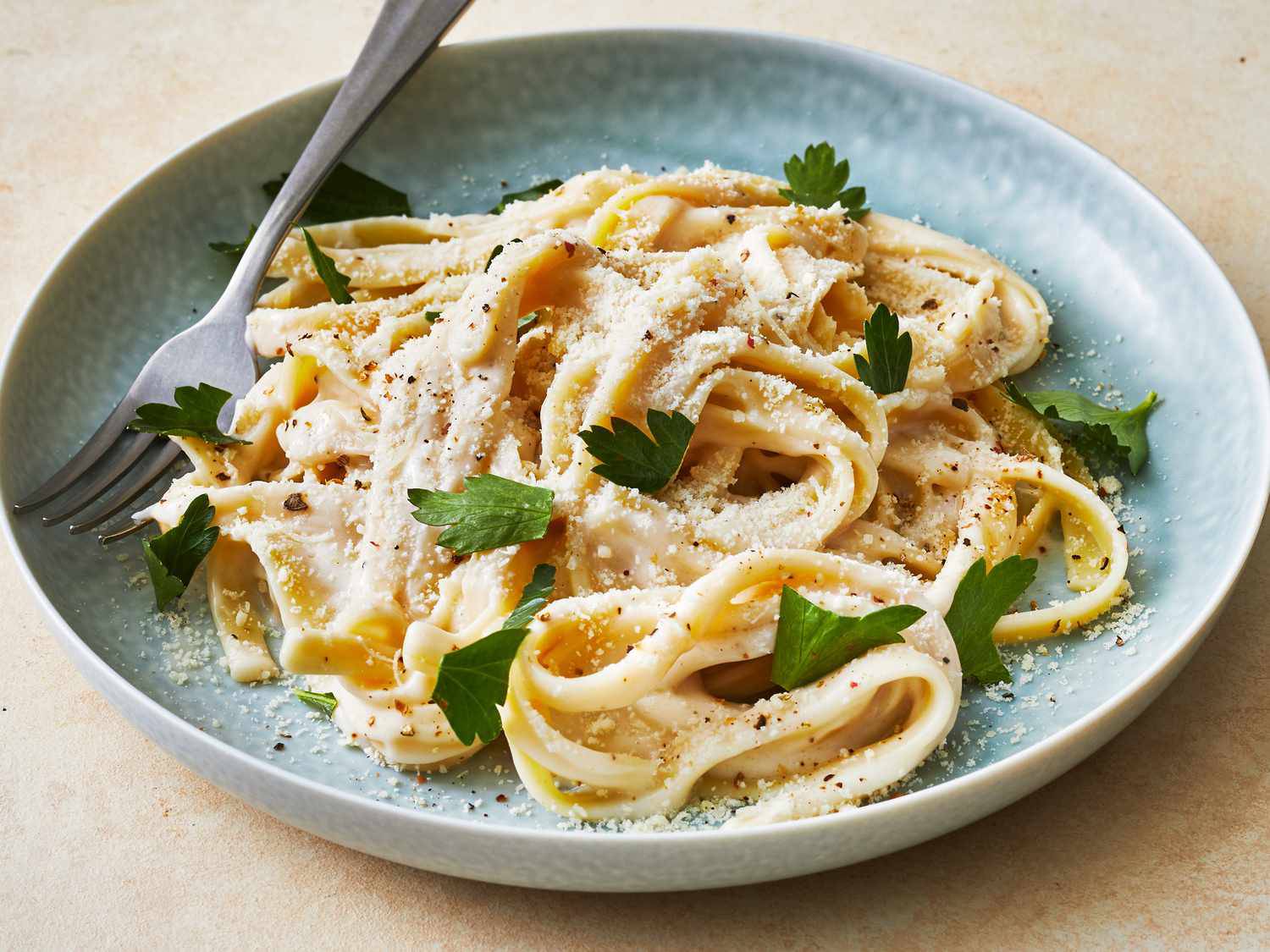 Creamy fettuccine pasta topped with grated Parmesan and fresh parsley, served on a light blue plate with a fork.