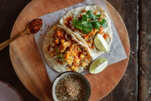 Two tacos filled with corn, guacamole, and toppings on a wooden platter, accompanied by a bowl of spices and lime wedges.
