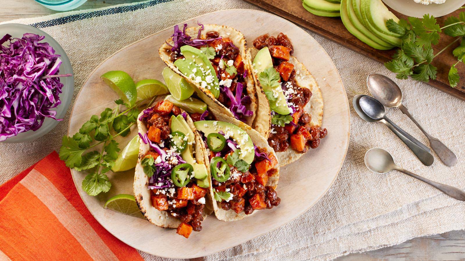 Three colorful tacos with black beans, sweet potatoes, avocado, and purple cabbage, garnished with lime and cilantro, on a wooden platter.