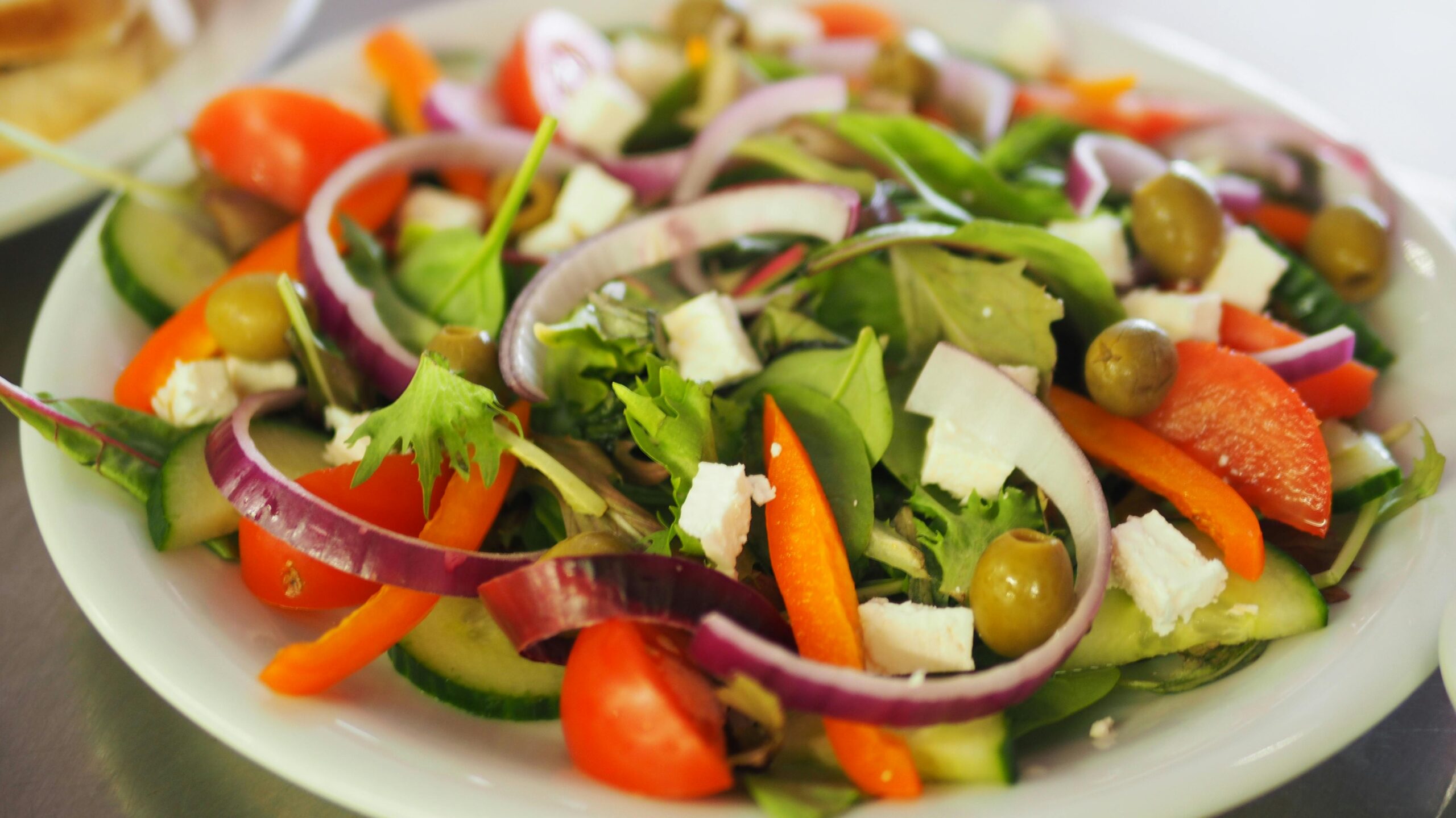 A vibrant salad featuring mixed greens, tomatoes, cucumbers, bell peppers, red onion, olives, and feta cheese on a white plate.