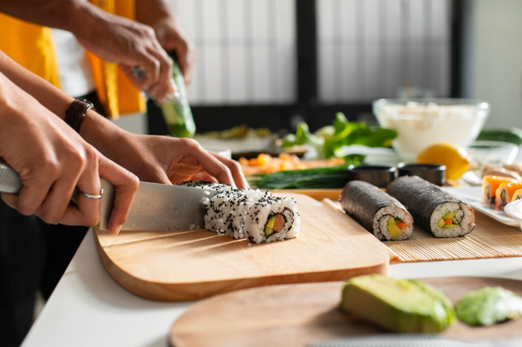 Two hands slicing sushi rolls on a wooden board, surrounded by fresh ingredients like cucumber, avocado, and sushi rice.