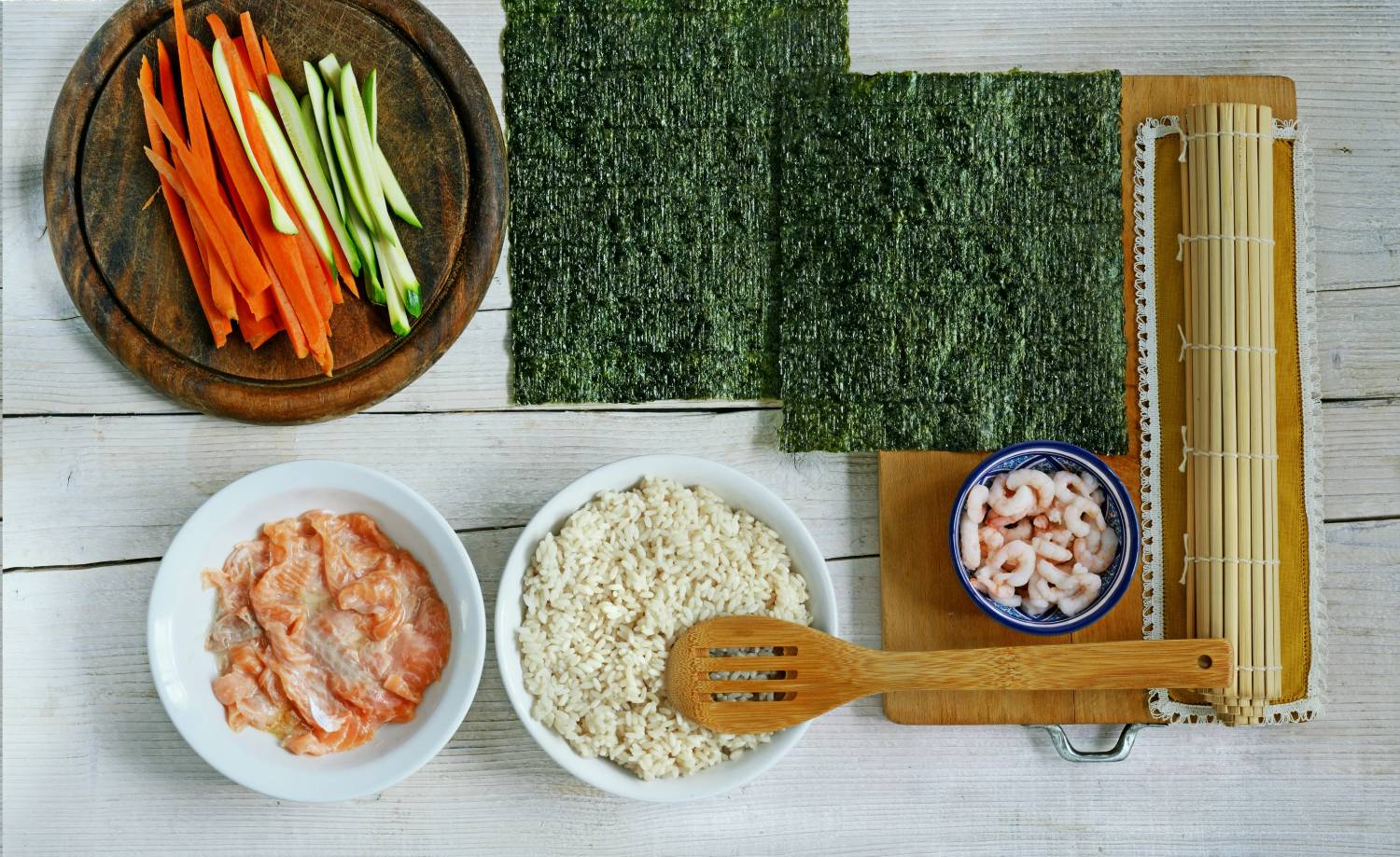 Ingredients for sushi preparation: sliced carrots, cucumbers, salmon, shrimp, rice, seaweed sheets, and a bamboo mat.