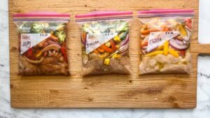 Three labeled ziplock bags on a wooden cutting board, containing marinated chicken and vegetables for Asian, Italian, and Fajita dishes.