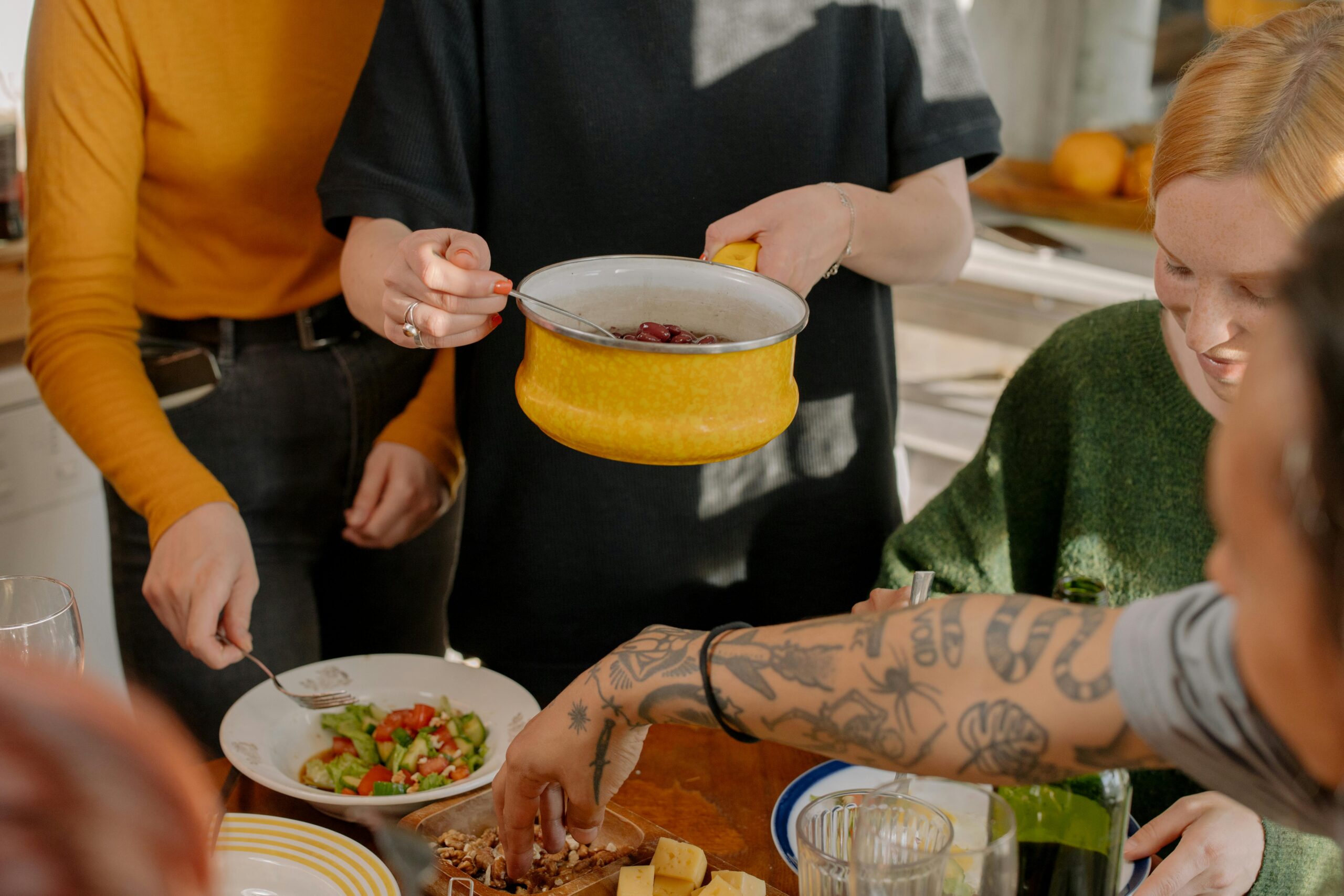 A group of people preparing a meal together, with a focus on a person holding a yellow pot and others serving salad and snacks.