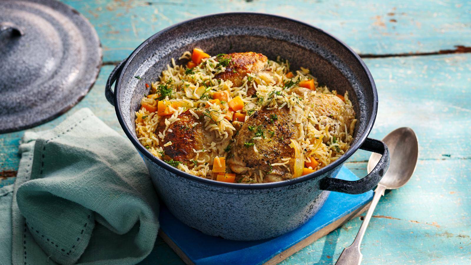 A pot filled with chicken and rice, garnished with herbs and chunks of orange vegetables, sits on a rustic teal wooden surface.