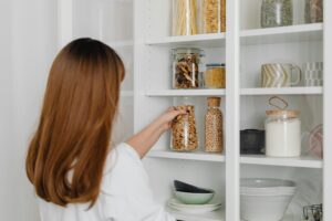 A woman with long brown hair organizes glass jars filled with food items on white shelves in a modern kitchen pantry.