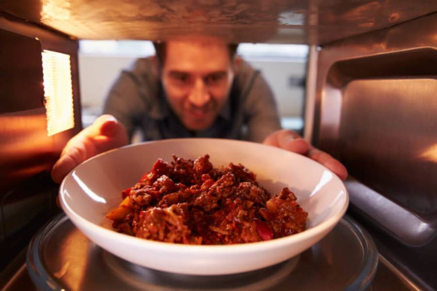 A person reaches into a microwave to retrieve a bowl of savory meat and vegetable stew.
