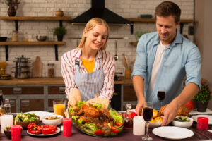 A festive table is set with a whole roasted chicken on greens, candles, salads, and drinks, with two people preparing for the meal.