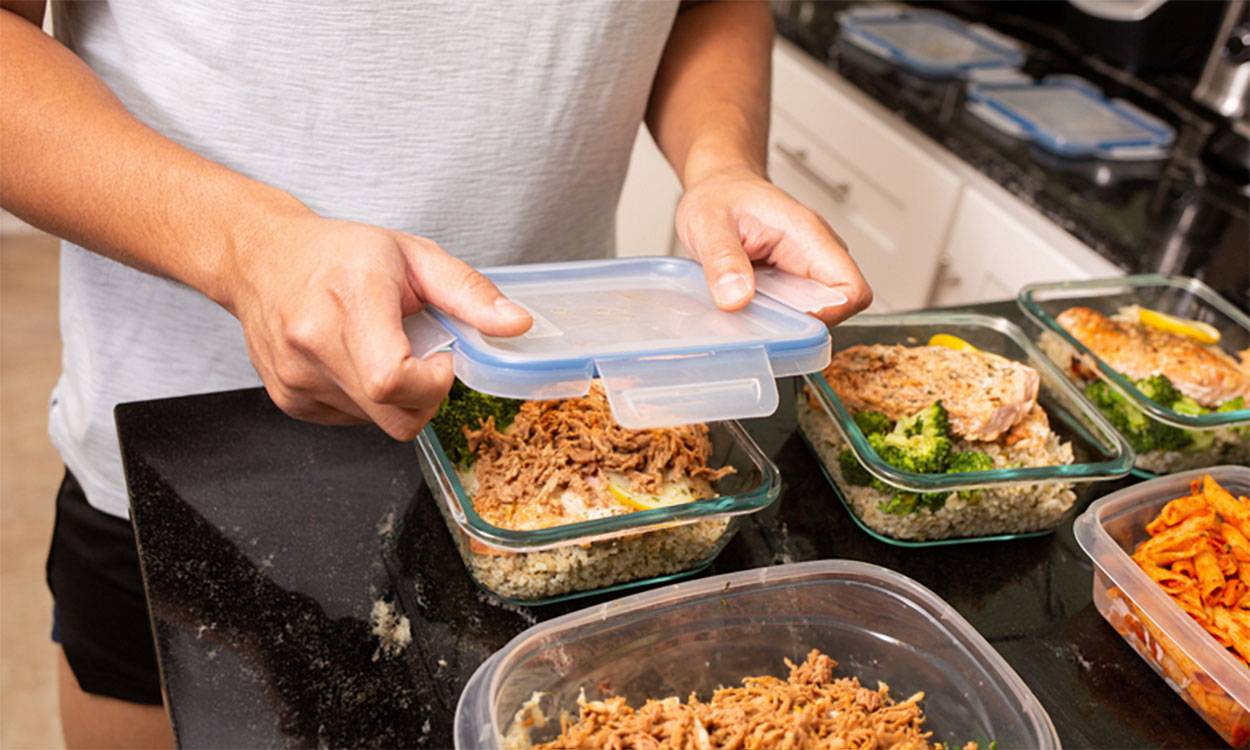 A person seals a meal prep container filled with pulled meat, broccoli, and rice, surrounded by other meal containers on a countertop.
