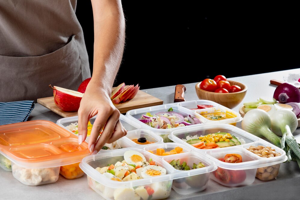 A hand reaches to organize colorful meal prep containers filled with vegetables, eggs, and fruits on a kitchen countertop.