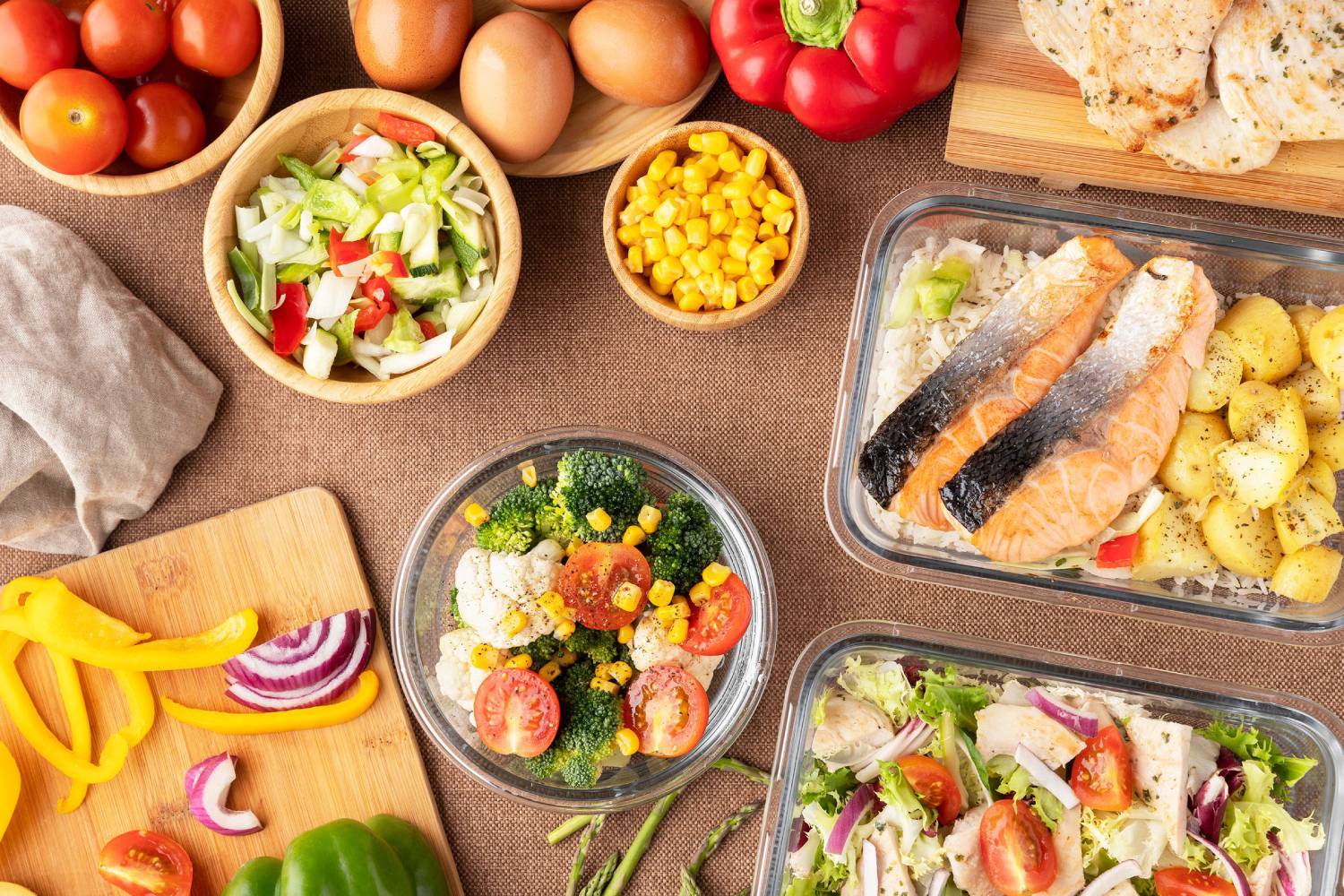 An assortment of fresh vegetables, cooked salmon, and meal prep containers filled with healthy dishes, alongside eggs and colorful bell peppers.