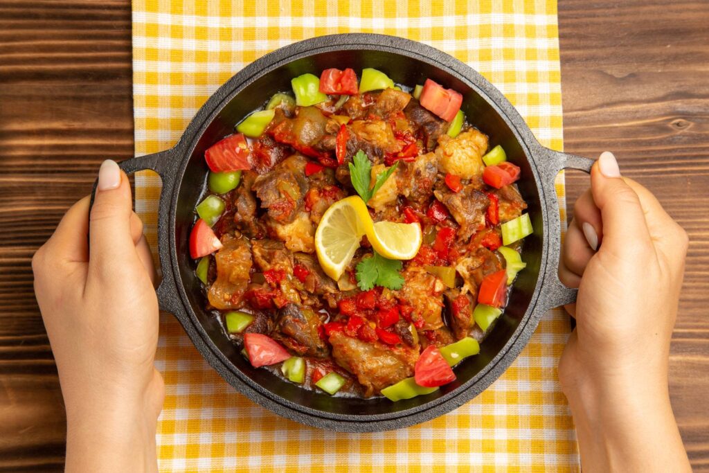 A person holds a sizzling dish of richly seasoned meat, colorful peppers, and tomatoes, garnished with lemon and cilantro on a checkered cloth.