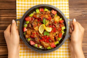A person holds a sizzling dish of richly seasoned meat, colorful peppers, and tomatoes, garnished with lemon and cilantro on a checkered cloth.