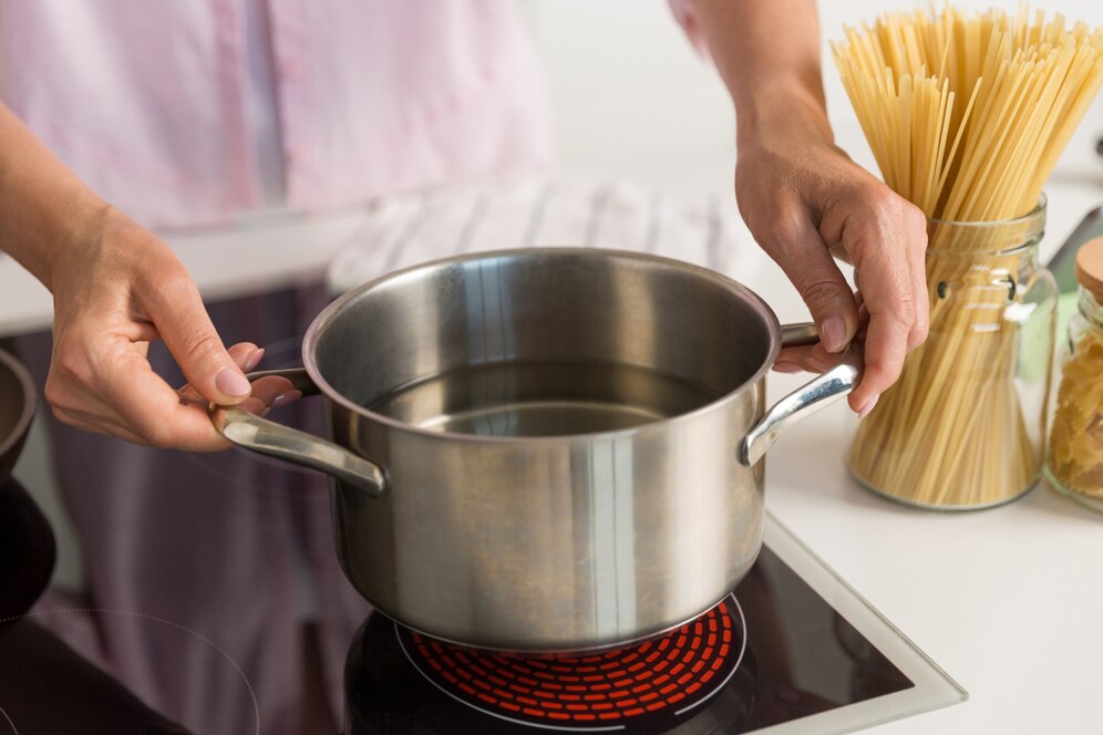A person holds a pot of water on a stovetop, preparing to cook pasta, with jars of spaghetti and ingredients nearby.