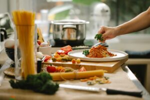A hand garnishes a plate of pasta with sauce and parsley amidst fresh ingredients and cooking utensils on a vibrant kitchen counter.