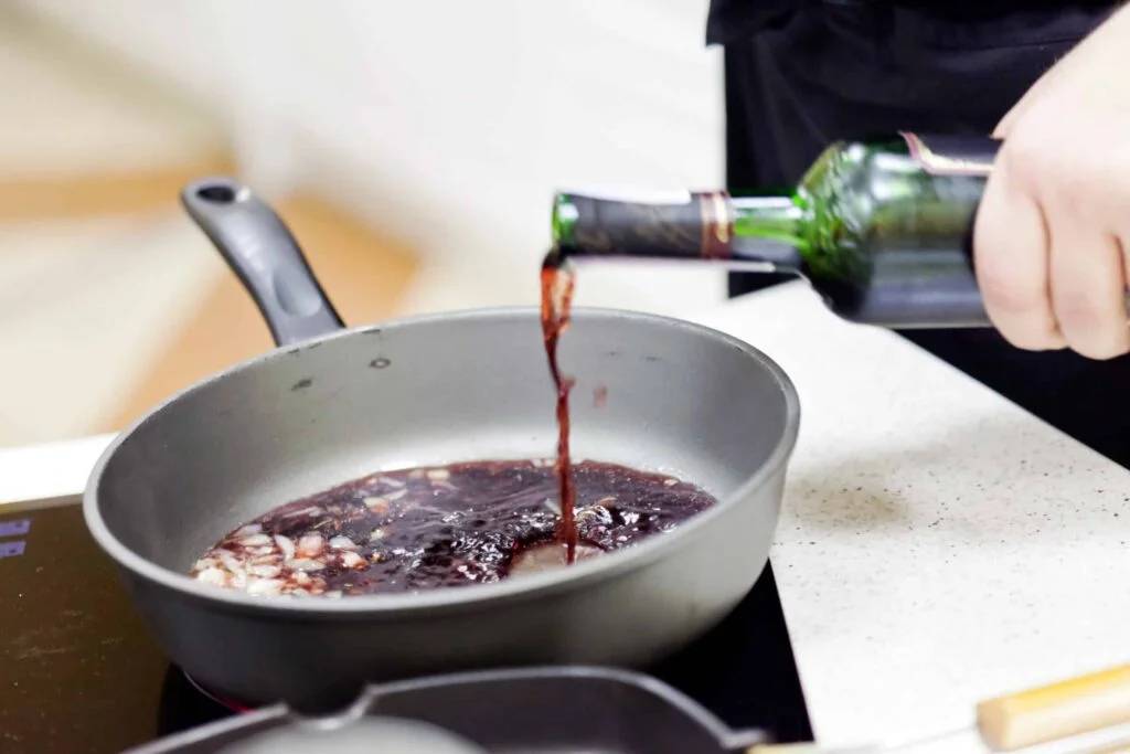 A hand pours red wine from a green bottle into a frying pan on a stove, mixing with ingredients in the pan.