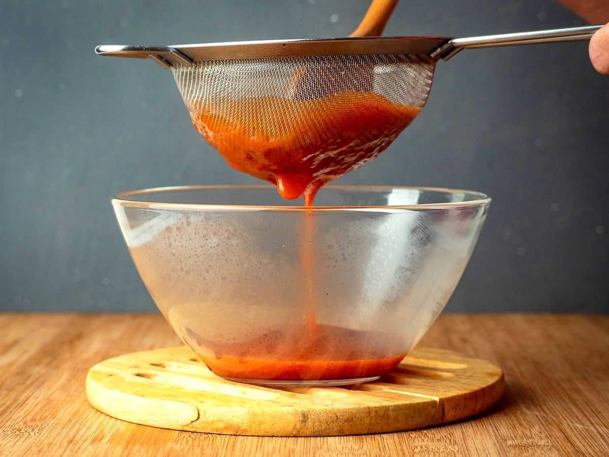 A hand holds a strainer over a clear bowl, pouring rich red sauce, with a wooden cutting board beneath.
