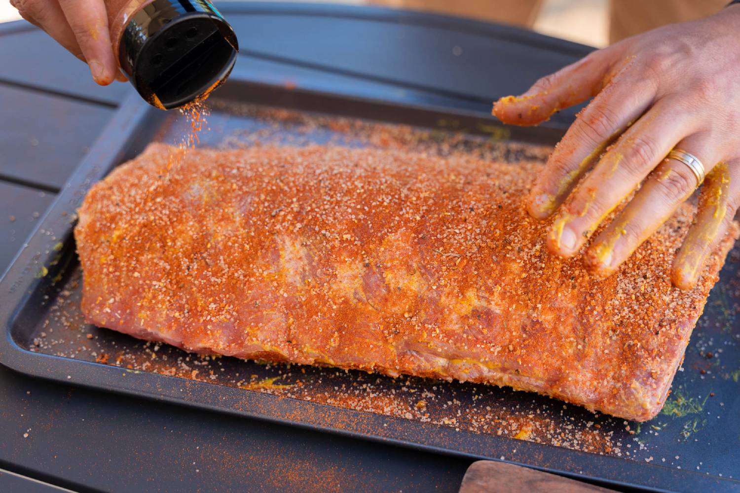 A hand sprinkles seasoning on a large piece of raw meat, covered in a blend of spices, on a black cooking tray.