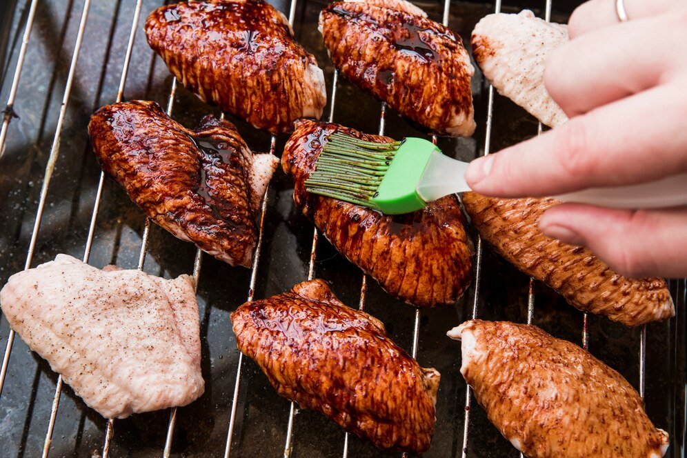 A hand brushes barbecue sauce onto grilled chicken wings on a rack, highlighting their glazed, caramelized finish.