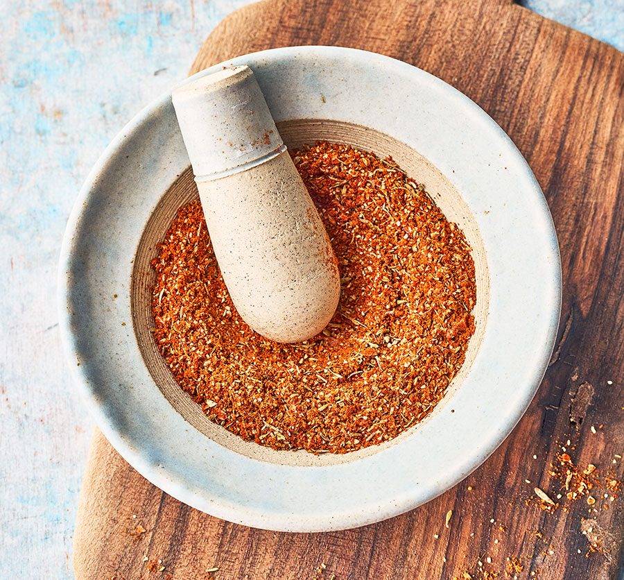 Close-up of a mortar and pestle filled with vibrant orange spice blend, set on a wooden surface against a blue backdrop.