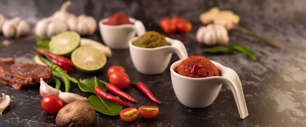 Three bowls of vibrant spice pastes surrounded by fresh herbs, garlic, chilies, tomatoes, and lime on a dark textured surface.
