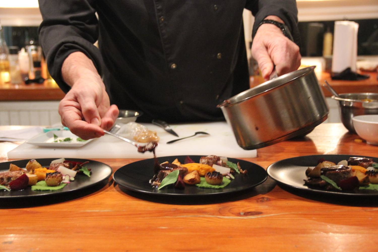 A chef plating three elegant dishes, drizzling sauce from a saucepan onto colorful vegetables and meats on black plates.