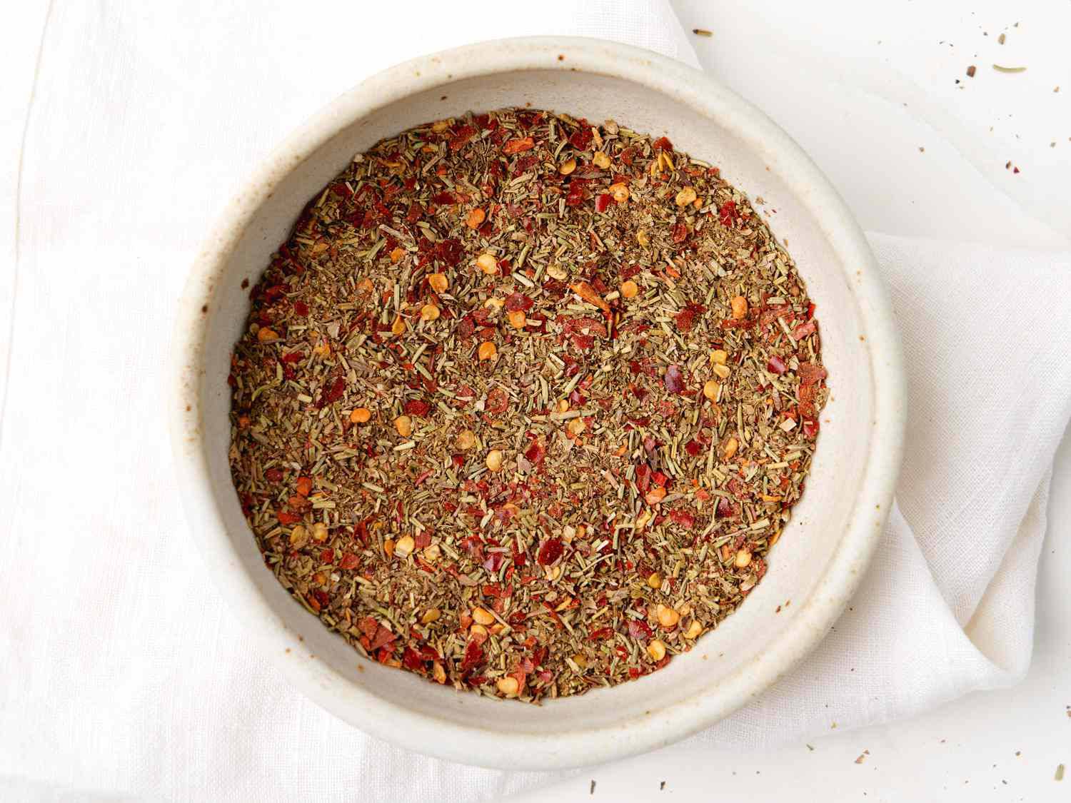 Close-up of a white bowl filled with a colorful blend of dried herbs and spices, resting on a white cloth background.