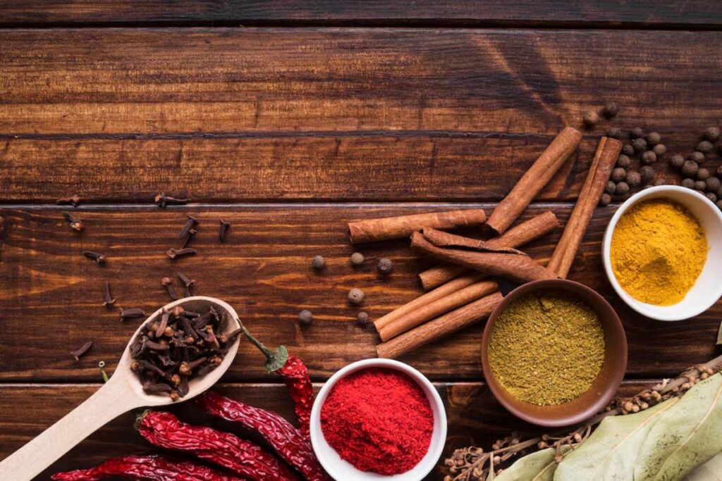 An assortment of spices on a wooden surface, including dried red chilies, cloves, cinnamon sticks, and various spice powders in bowls.