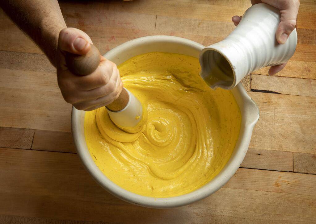 A person's hand holding a pestle mixes yellow batter in a white bowl, while another hand pours liquid from a ceramic container.