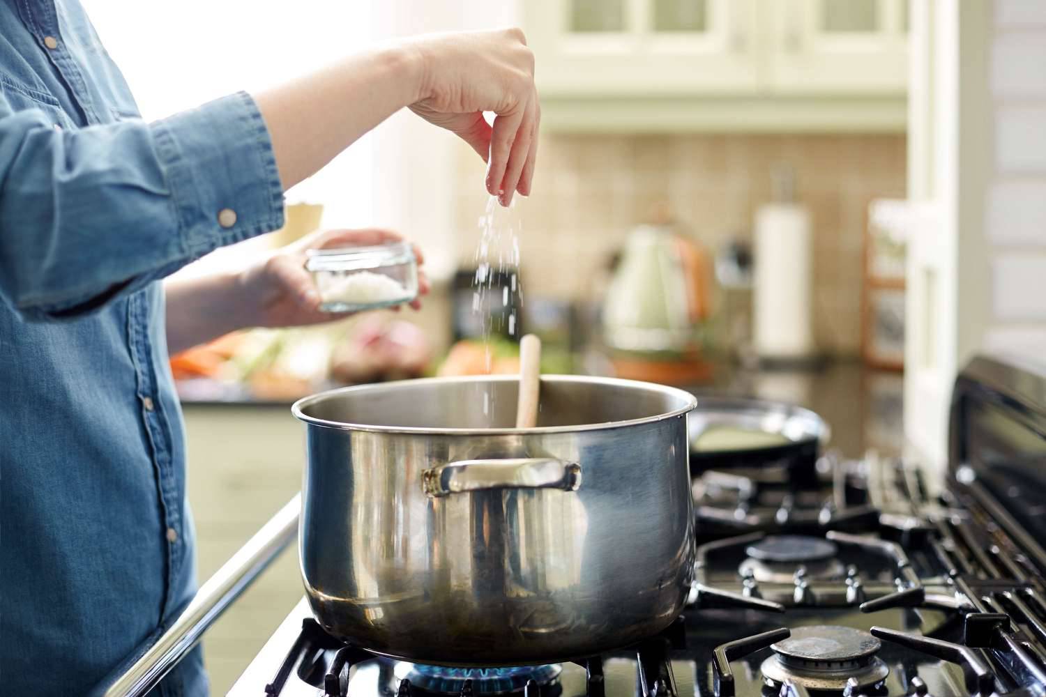 A person in a denim shirt adds salt from a jar into a pot on the stovetop, surrounded by colorful kitchen items in the background.