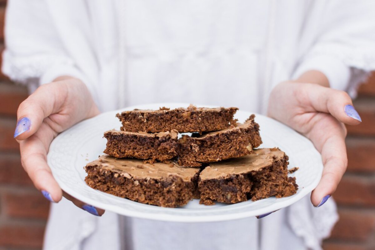 A person holding a white plate with stacked, frosted chocolate brownies, standing against a brick wall background.