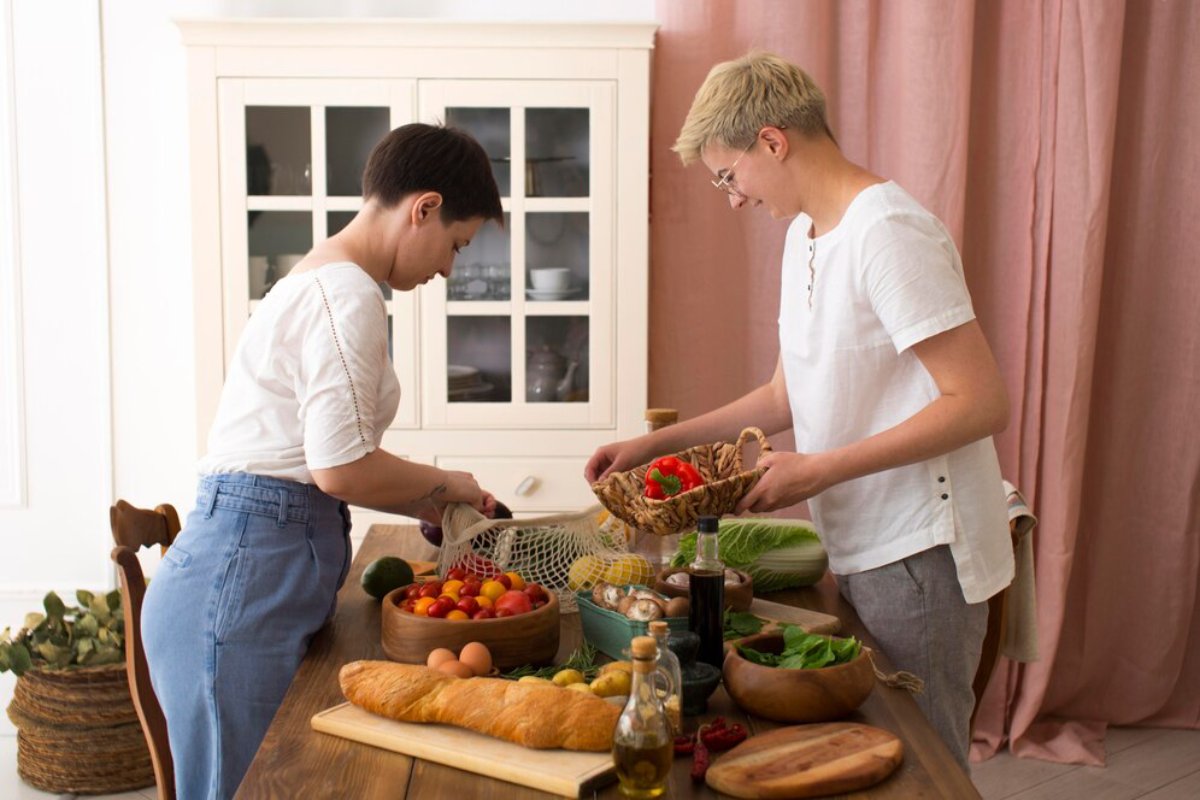 Two individuals prepare food in a bright kitchen, surrounded by fresh vegetables, fruits, and baked goods on a wooden table.
