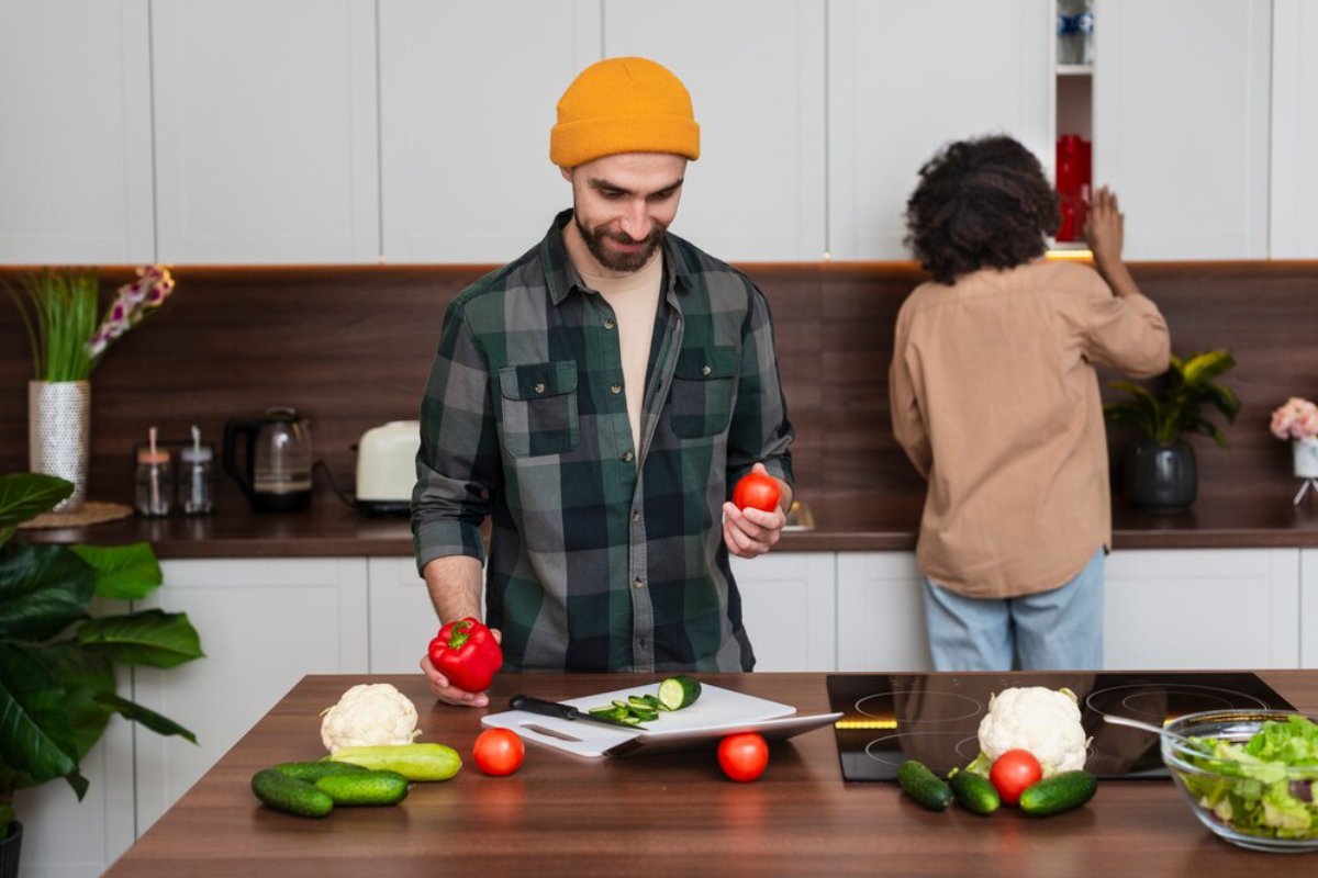 A person wearing a yellow beanie holds a red bell pepper and tomato, surrounded by various vegetables in a modern kitchen.