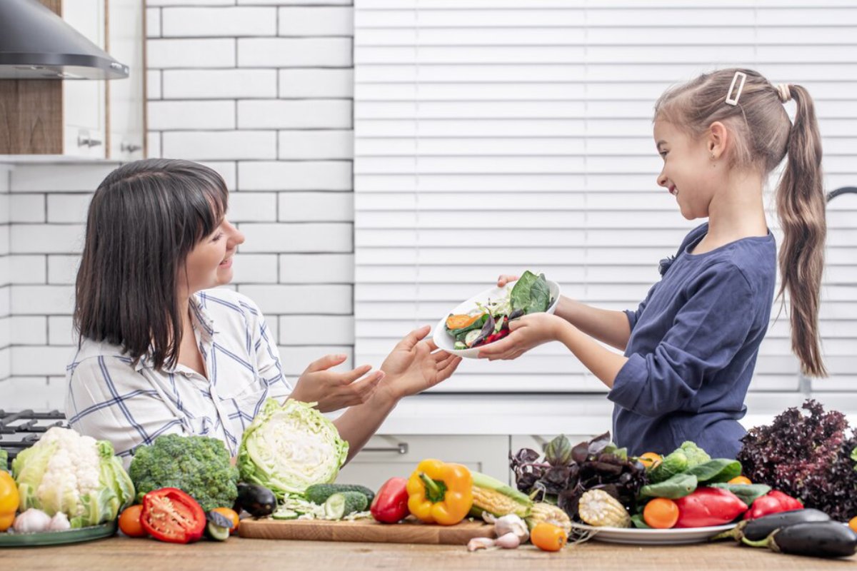 A child offers a plate of fresh vegetables to an adult in a bright kitchen filled with assorted colorful produce on the table.