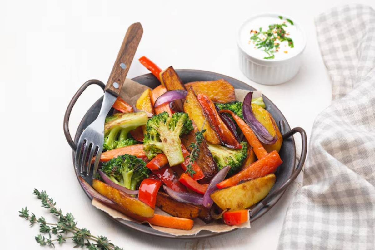 A pan of roasted vegetables including broccoli, carrots, peppers, and potatoes, served with a side of herb yogurt sauce.