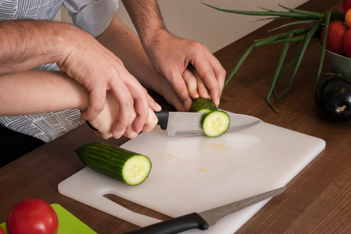 Two hands collaboratively slice a cucumber on a cutting board, with tomatoes and green onions in the background.