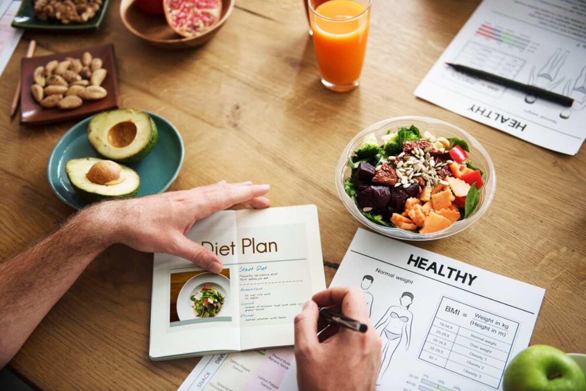 A person writing in a diet plan booklet beside a fresh salad, healthy snacks, and nutrition sheets on a wooden table.