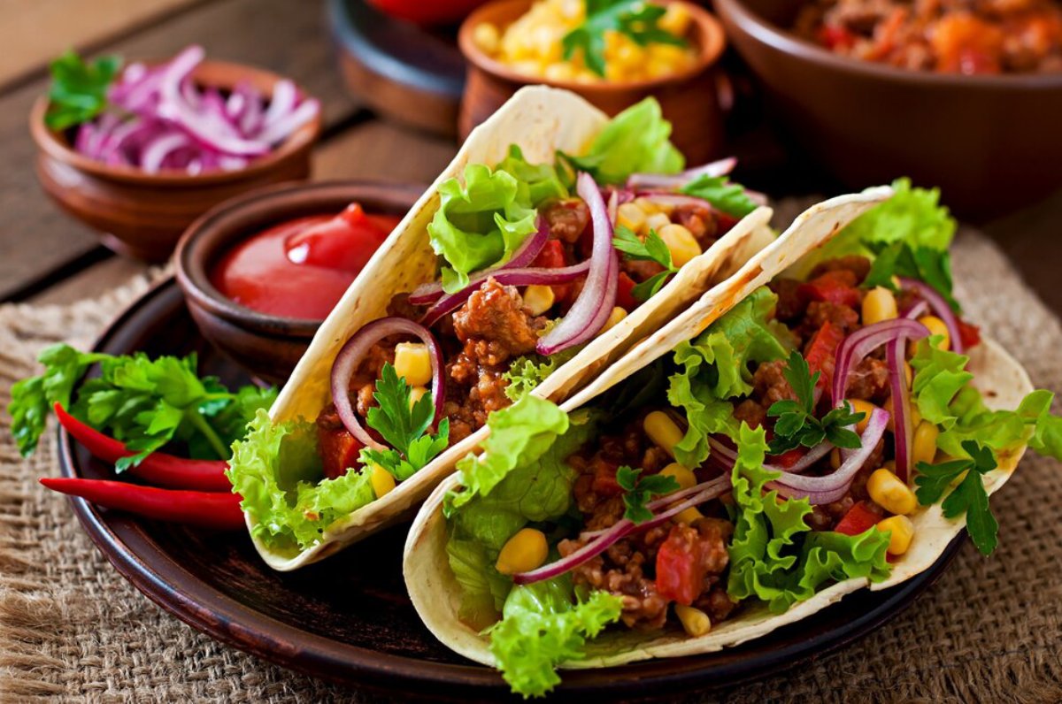 Two loaded tacos filled with ground meat, lettuce, corn, tomatoes, and red onions, served with dipping sauces on a rustic plate.